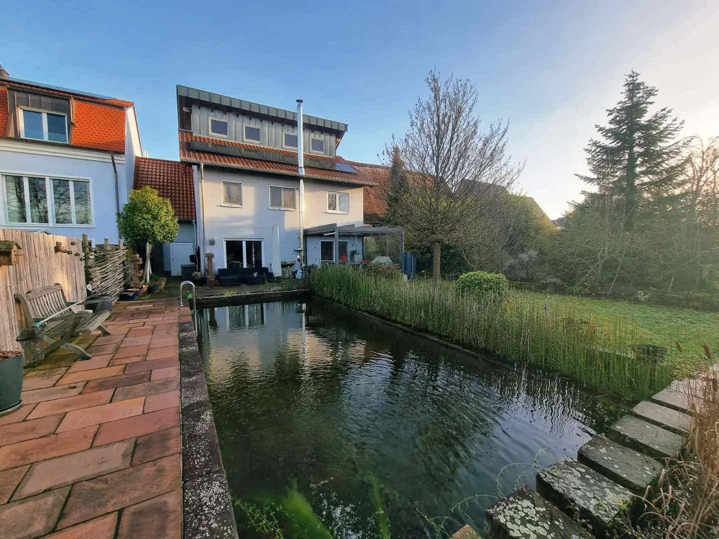 Backyard view of a two-story house with a pond, patio, and green lawn under a blue sky.
