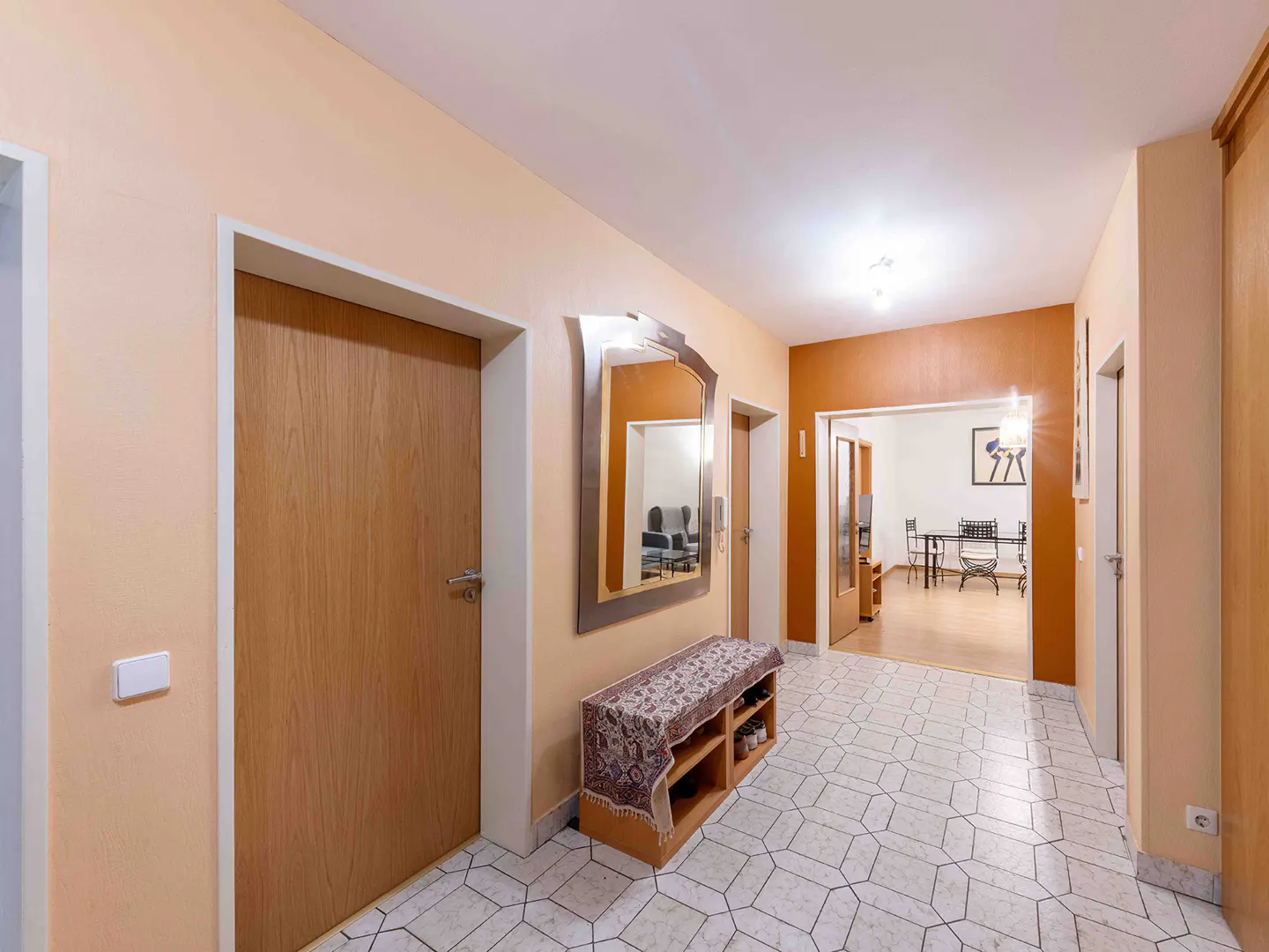 Hallway with peach walls, white tile floor, wooden doors, mirror, and shoe rack. Dining room visible in background.