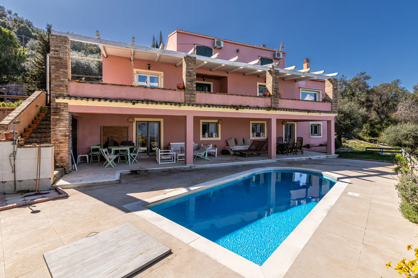 Two-story pink house with a blue pool in the foreground on a sunny day. Outdoor furniture is visible on the patio.