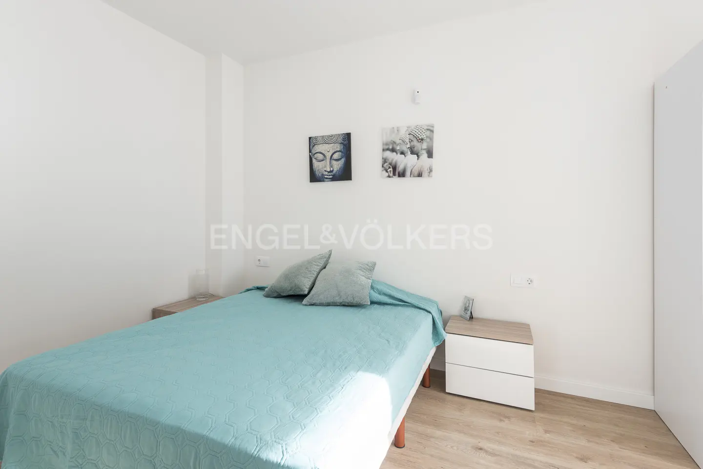 Bright bedroom with a blue bedspread, two pillows, and two framed pictures of Buddha on the white wall. A nightstand is on each side of the bed.