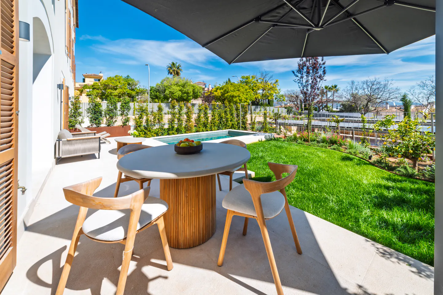 Outdoor patio with round table, chairs, and umbrella. A pool and green lawn are in the background under a blue sky.