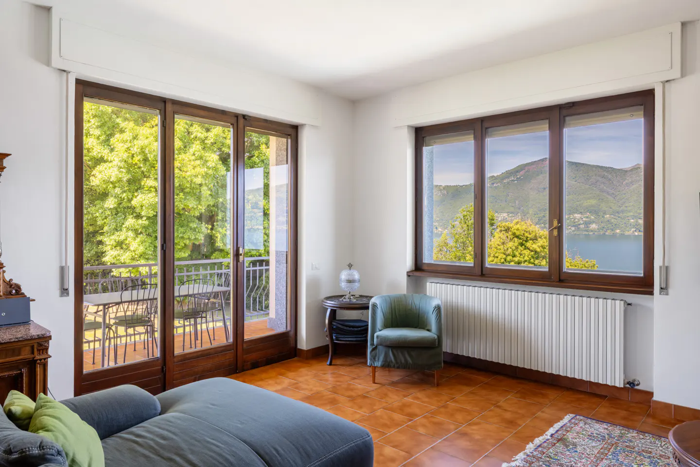 Living room with terracotta floors, a green armchair, and large windows overlooking a lake and mountains.