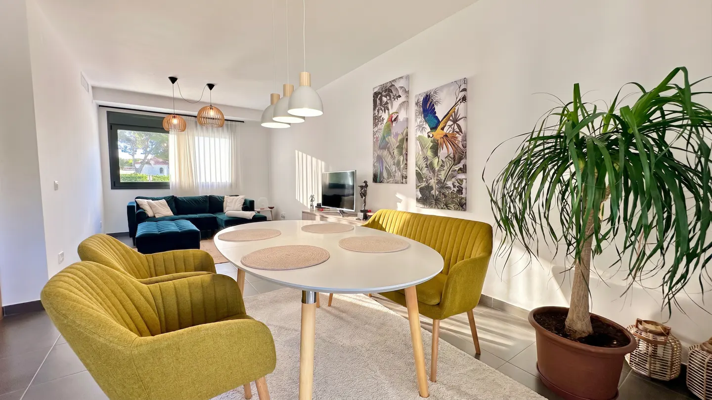 Bright living room with a white table, four yellow chairs, and a green velvet sofa. Parrot art hangs on the wall.