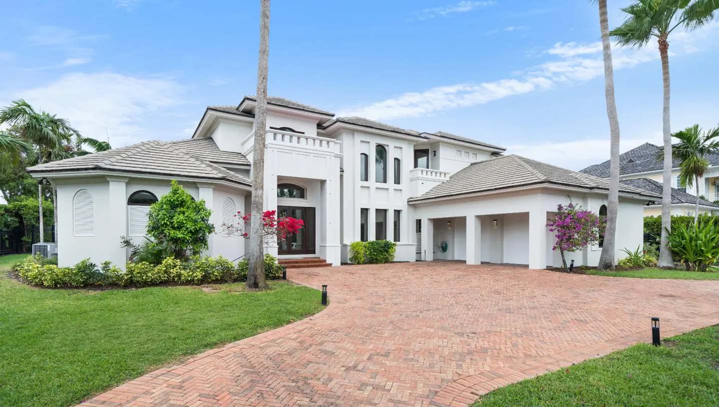 Exterior view of a large, white two-story house with a brick driveway, green lawn, and palm trees under a blue sky.