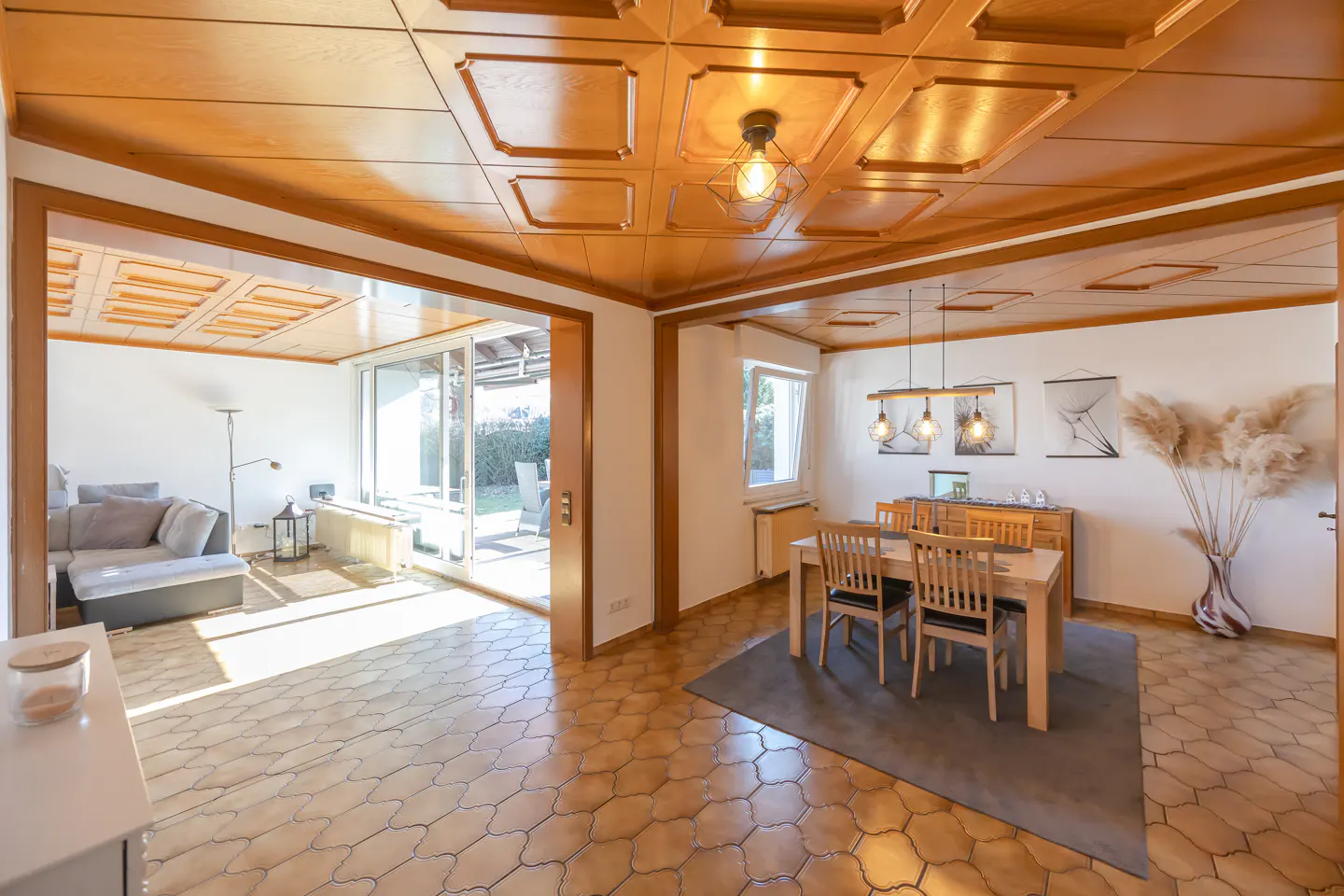 Open-concept living and dining room with wood-paneled ceiling and tile floor. Dining table with four chairs and a sideboard. Living room with sofa and sliding glass doors.