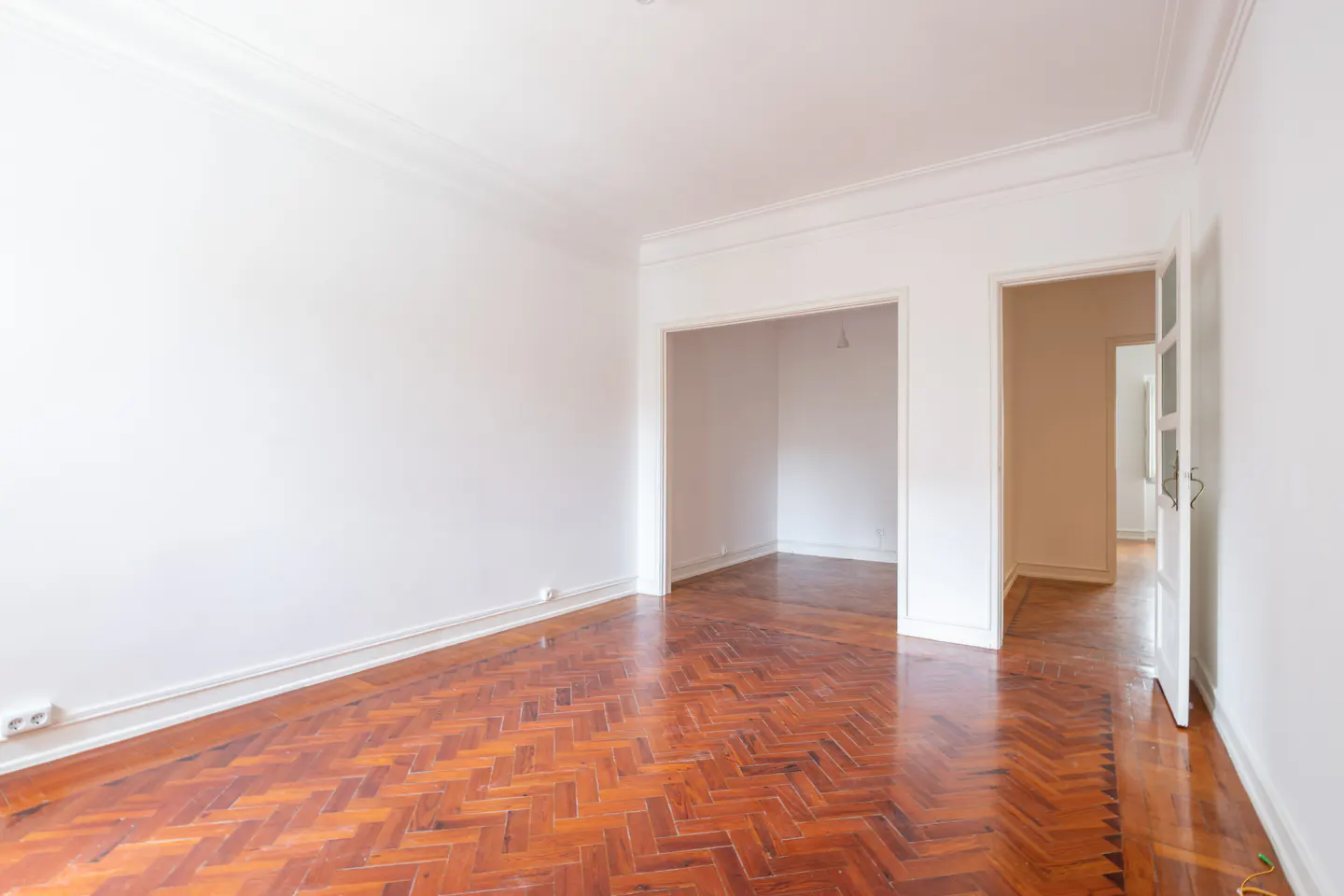 Empty room with white walls, herringbone wood floor, and doorways leading to other rooms.