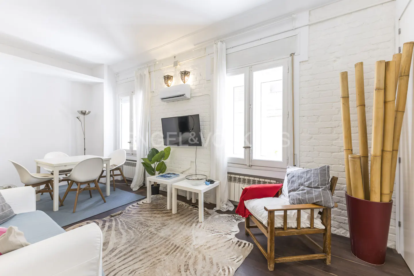Bright living room with white brick walls, a dining table, a TV, and a wooden chair next to a red vase with bamboo.