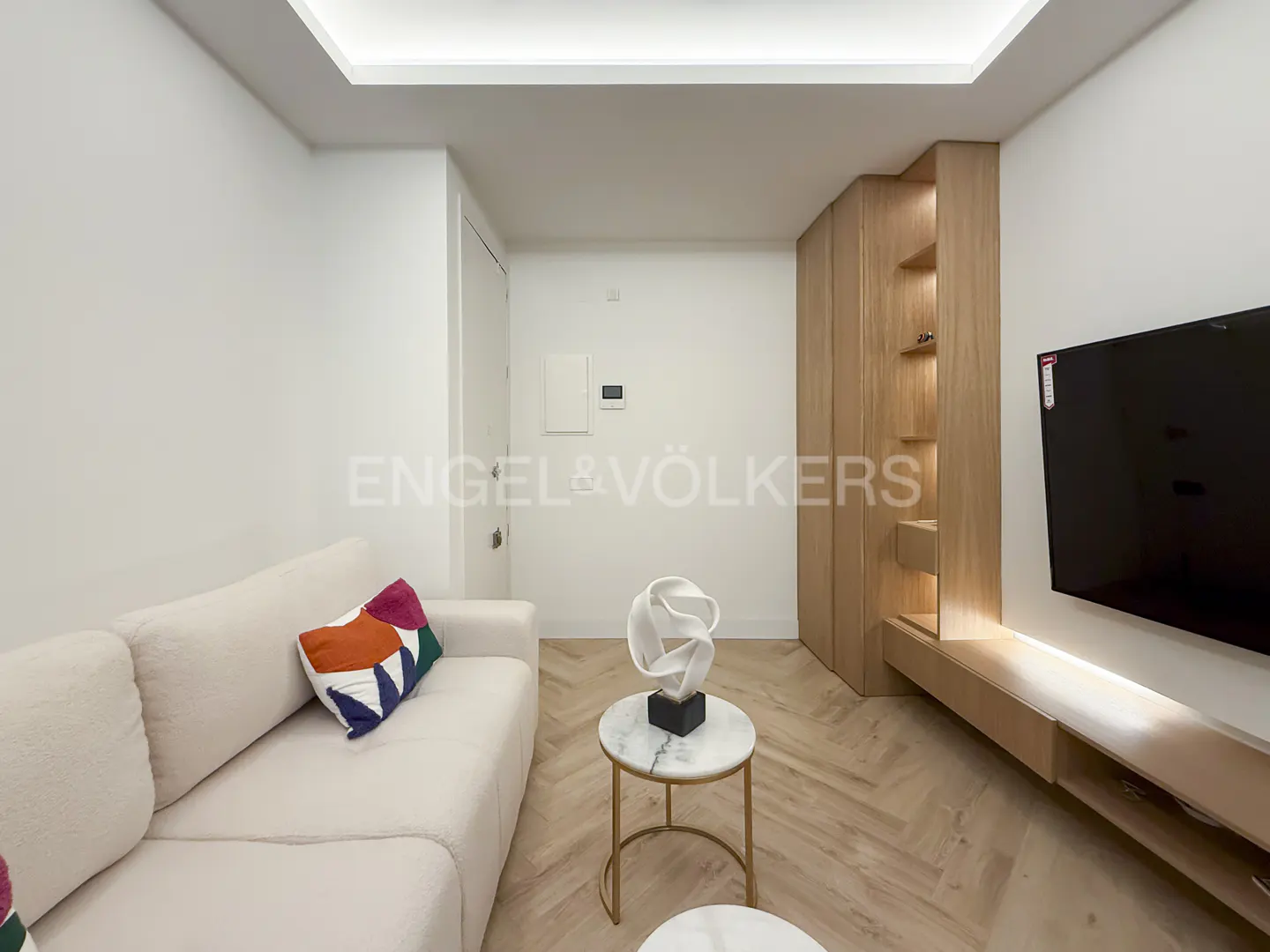 Living room with white walls, herringbone wood floors, and a cream sofa with colorful pillows. A modern sculpture sits on a marble-topped table.
