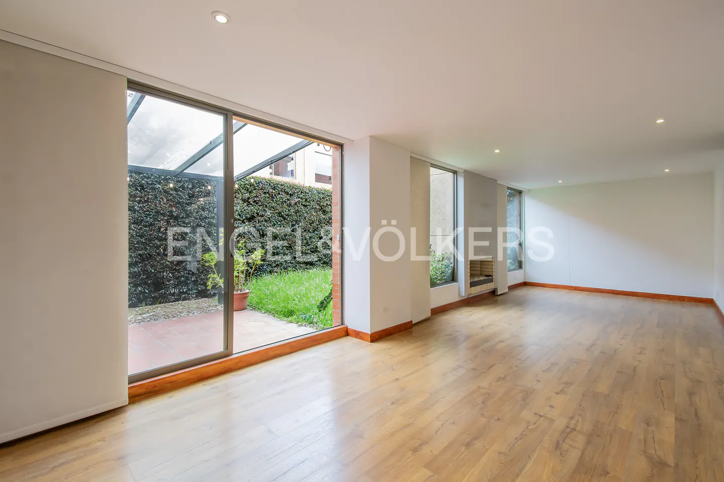 Bright, empty living room with hardwood floors and large windows overlooking a green, enclosed patio.