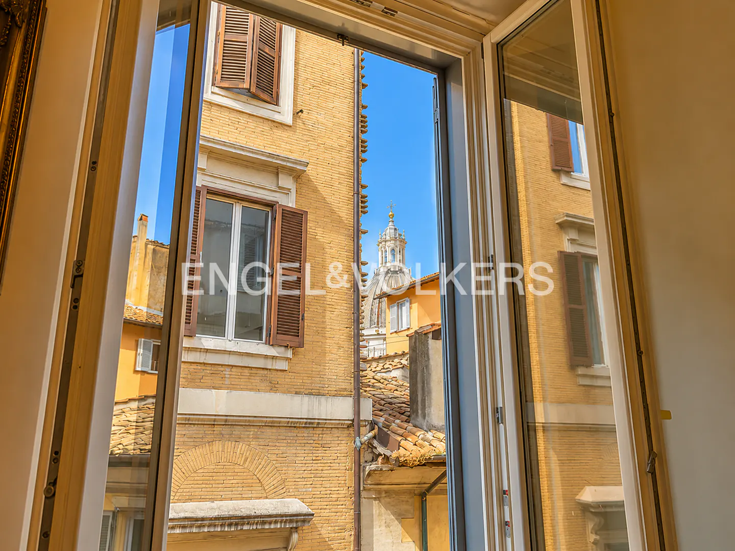 View from an open window of a yellow brick building with brown shutters and a church dome in the distance.