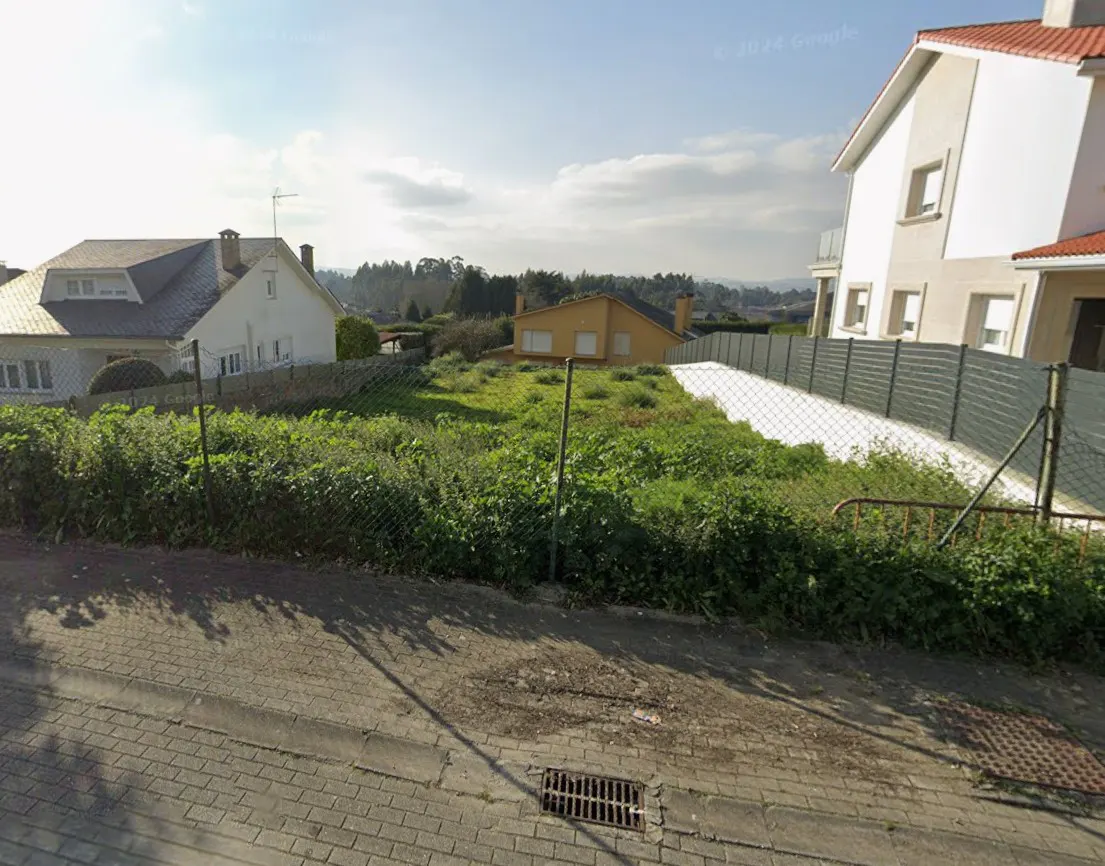 A vacant lot with green grass and weeds is fenced in between two houses on a sunny day.