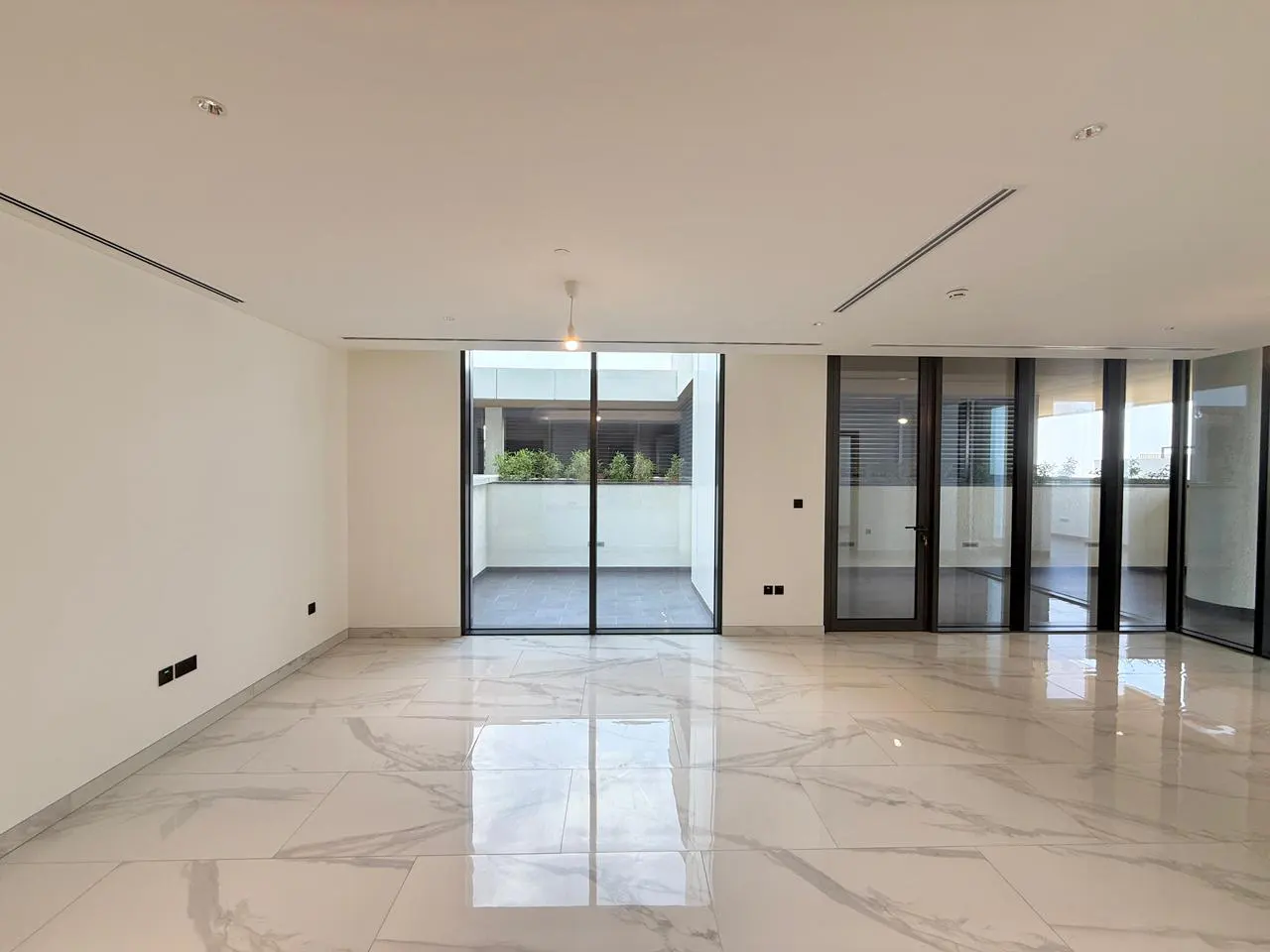 Empty, bright room with white walls and marble floors. Large glass doors lead to a balcony with greenery.