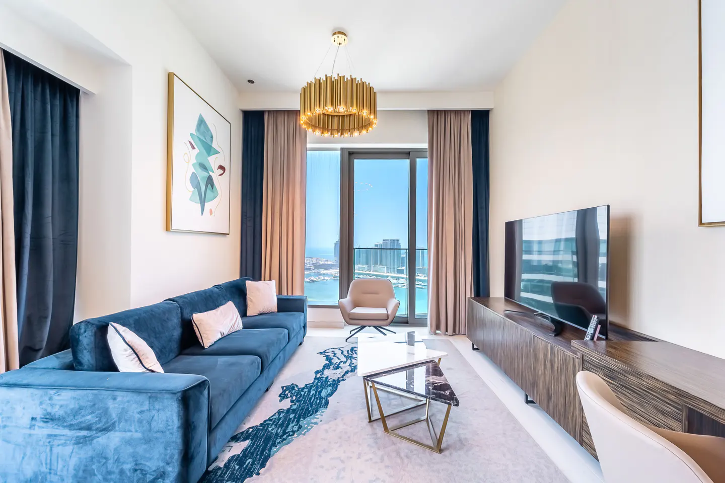 Modern living room with a blue velvet sofa, gold chandelier, TV console, and floor-to-ceiling windows overlooking the city.