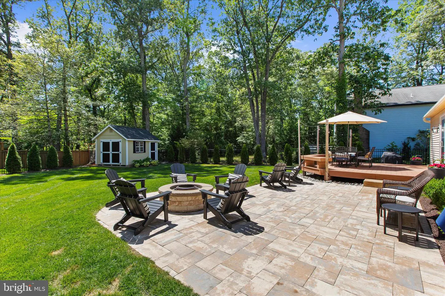 A backyard with a stone patio, fire pit with chairs, a deck with a table and umbrella, and a shed.