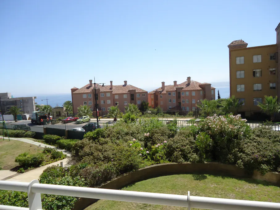 View from a balcony overlooking a complex of peach-colored buildings with red tile roofs, green landscaping, and a glimpse of the ocean.