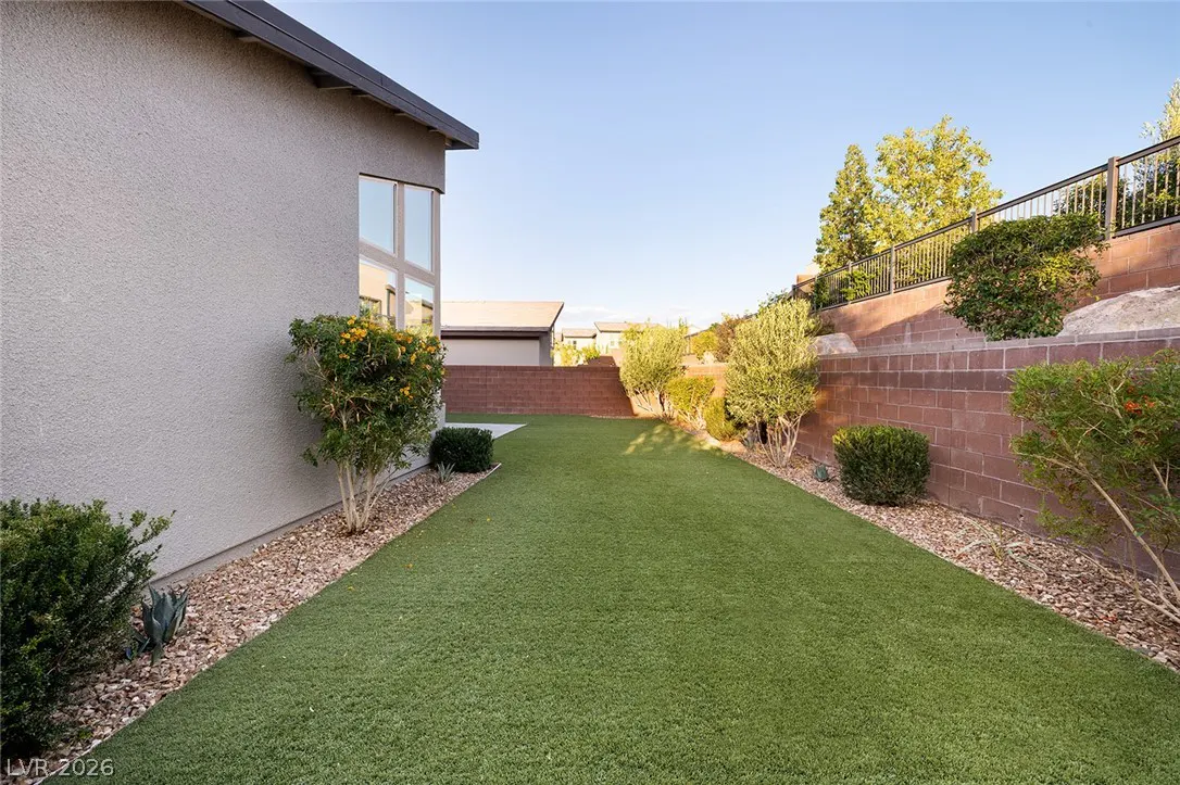 A backyard with green grass, a gray house, and a brick wall. Bushes and trees line the wall. The sky is blue.