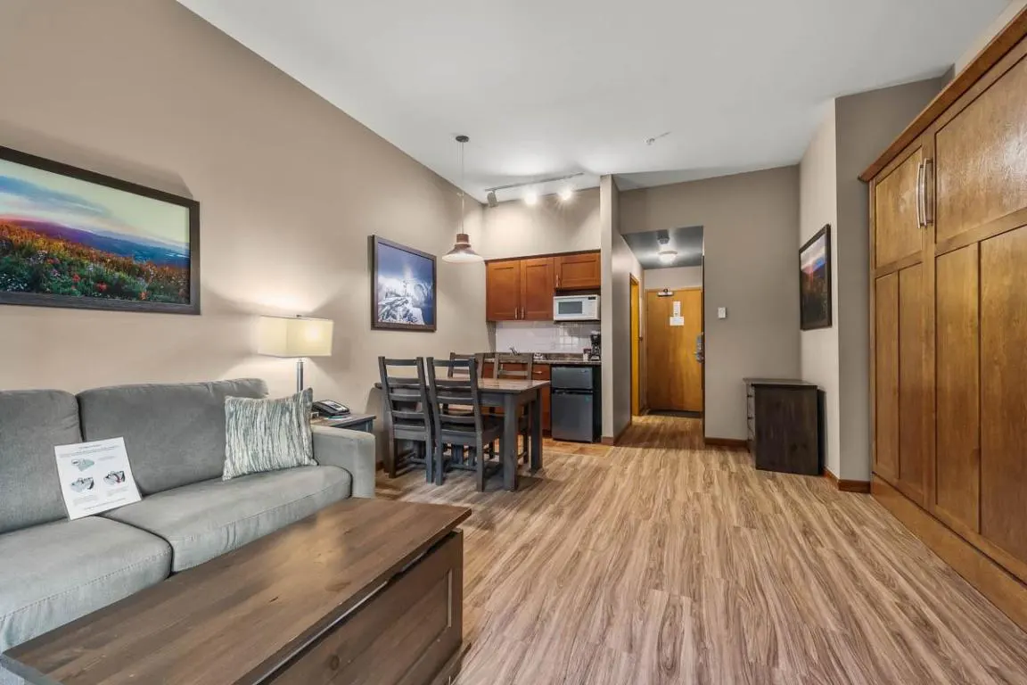 Interior view of a hotel suite with a gray sofa, wood floors, a dining table, and a kitchenette. Artwork hangs on the beige walls.