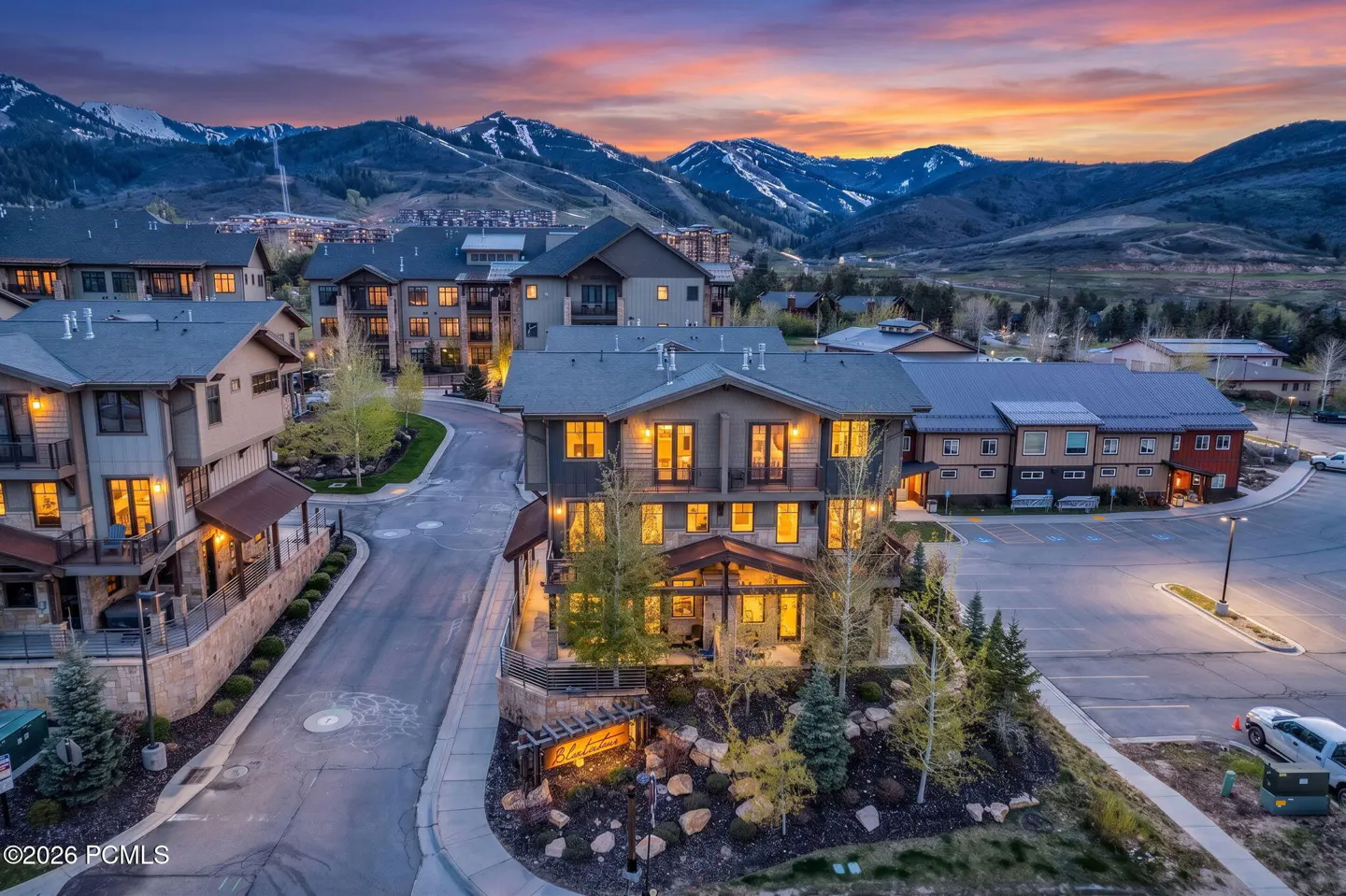 Aerial view of a mountain resort at dusk. Buildings with lit windows, parking lot, and snow-capped mountains in the background.