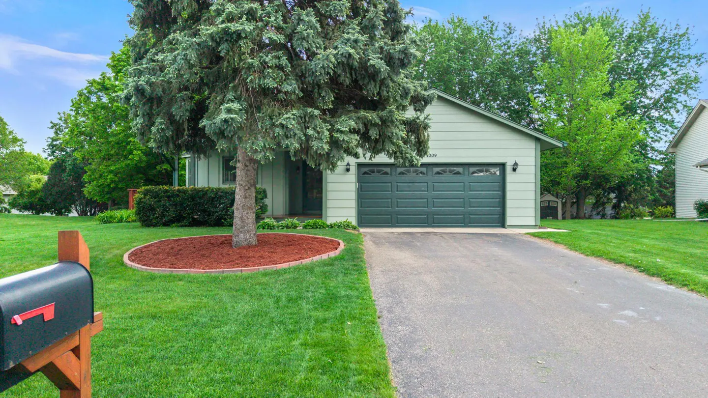 A light green single-story house with a dark green garage door and a large tree in the front yard.