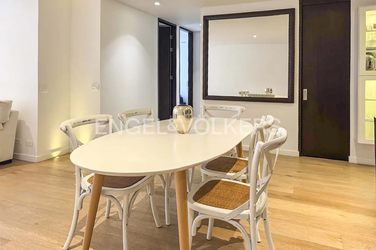 Modern dining area with an oval white table, four white cross-back chairs with woven seats, and a large wall mirror.