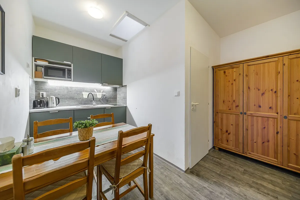 Small kitchen with dark green cabinets, a wooden dining table with chairs, and a large wooden wardrobe on the right.