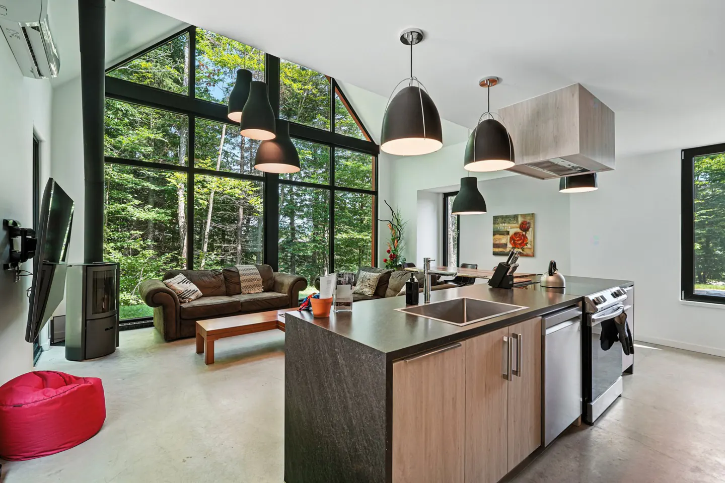 Open-concept living space with kitchen island, brown sofas, and large windows overlooking a forest. Black pendant lights hang above.