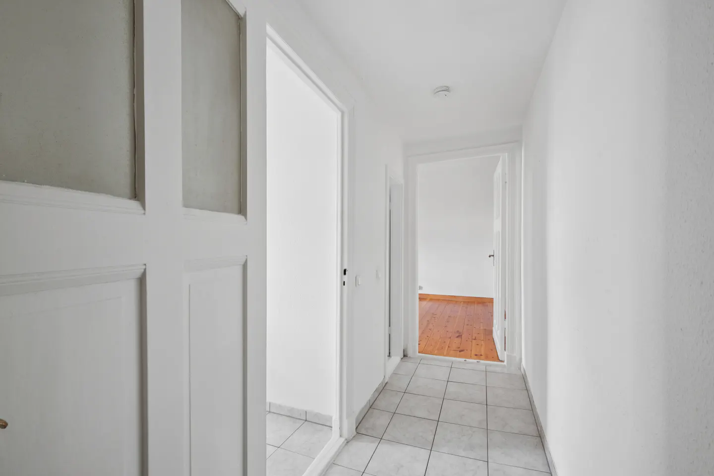 Bright hallway with white walls, tiled floor, and open doorways. A glimpse of hardwood flooring is visible in the room at the end of the hall.