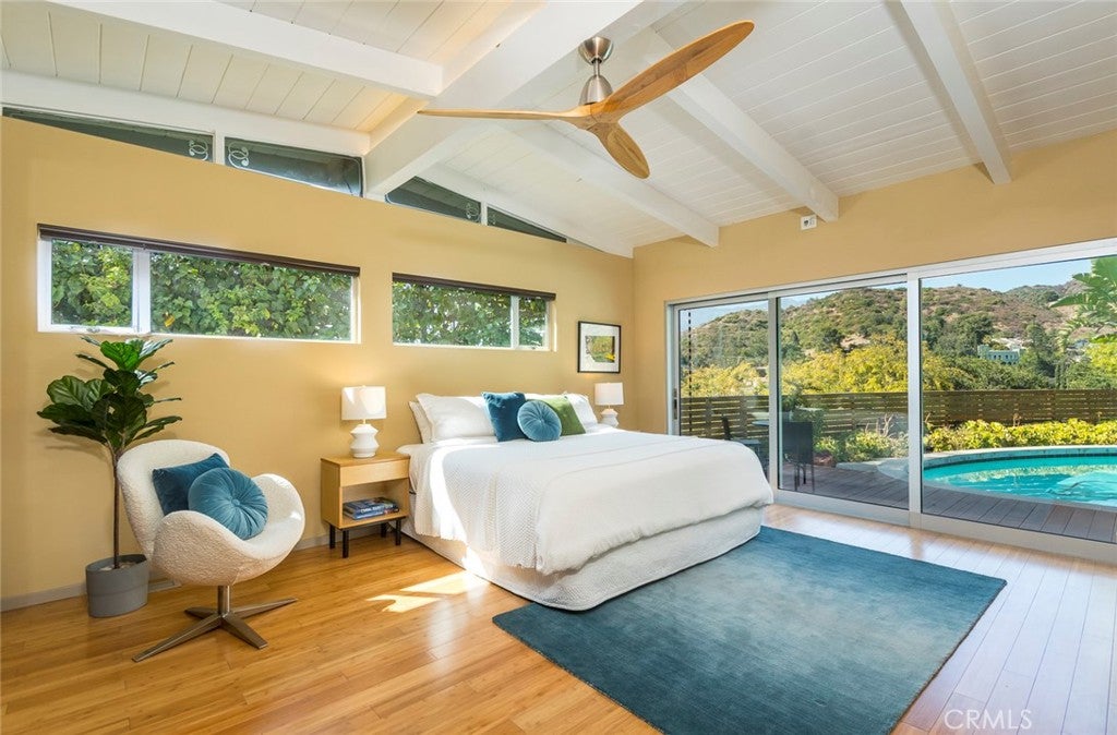 Modern bedroom with yellow walls, white vaulted ceiling, wood floors, bed, blue rug, and sliding doors to a pool deck. [AI-generated text]