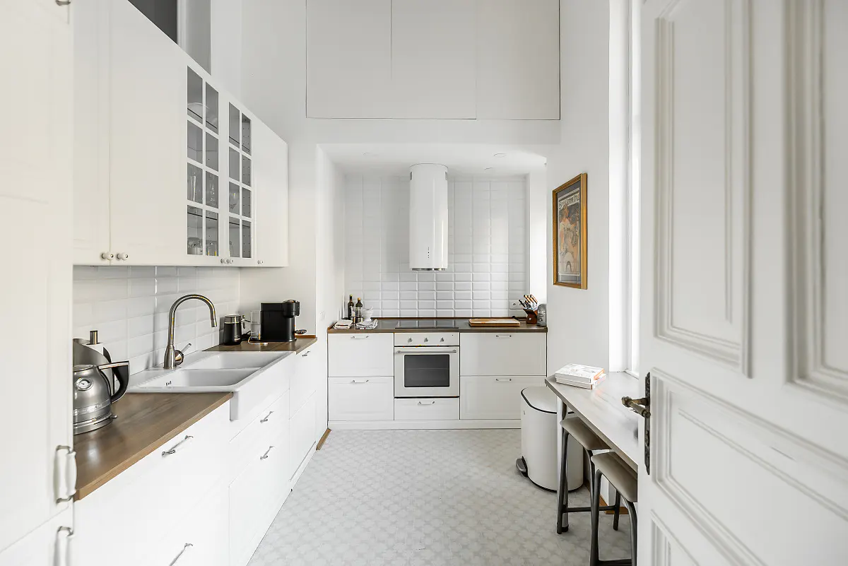 Bright, narrow kitchen with white cabinets, subway tile, and wood countertops. A small table and stools are near an open white door.