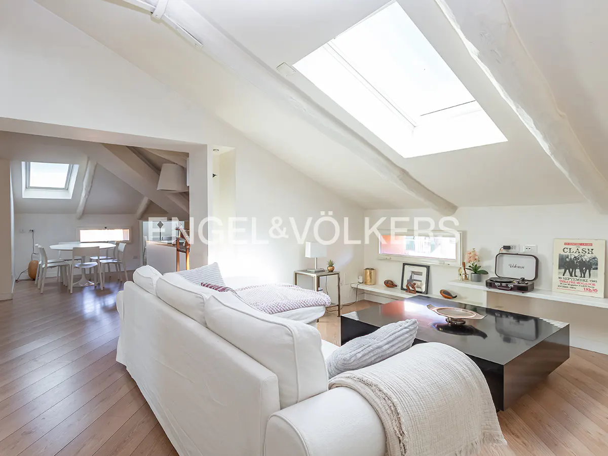 Bright attic living room with white sofa, black coffee table, skylight, and a dining area visible in the background.