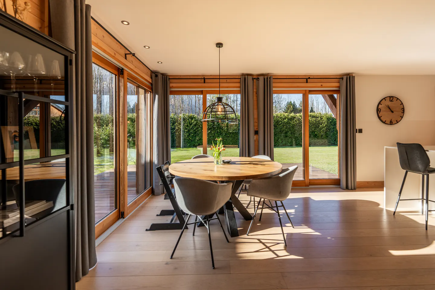 Bright dining room with wood floors, a round table with gray chairs, and large windows overlooking a green lawn.