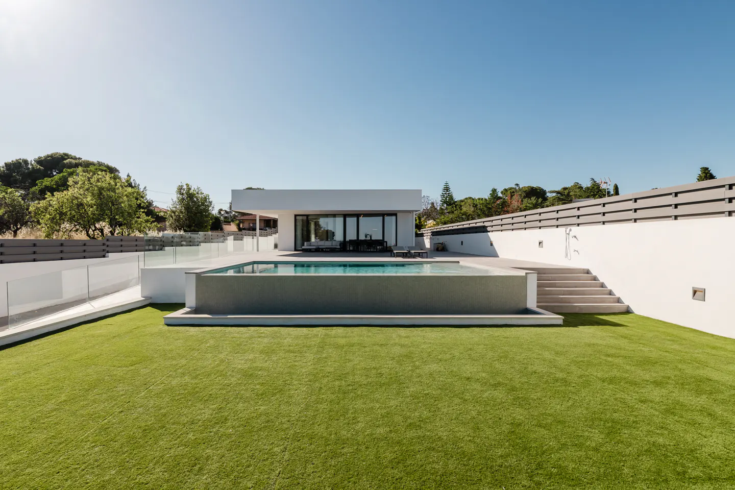 Modern white house with a rectangular pool, stone steps, and a green lawn under a clear blue sky.