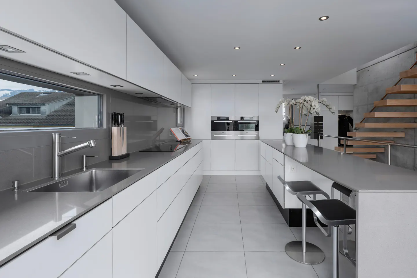 A modern kitchen with white cabinets, gray countertops, and stainless steel appliances. A window overlooks a town. A staircase is visible in the background.