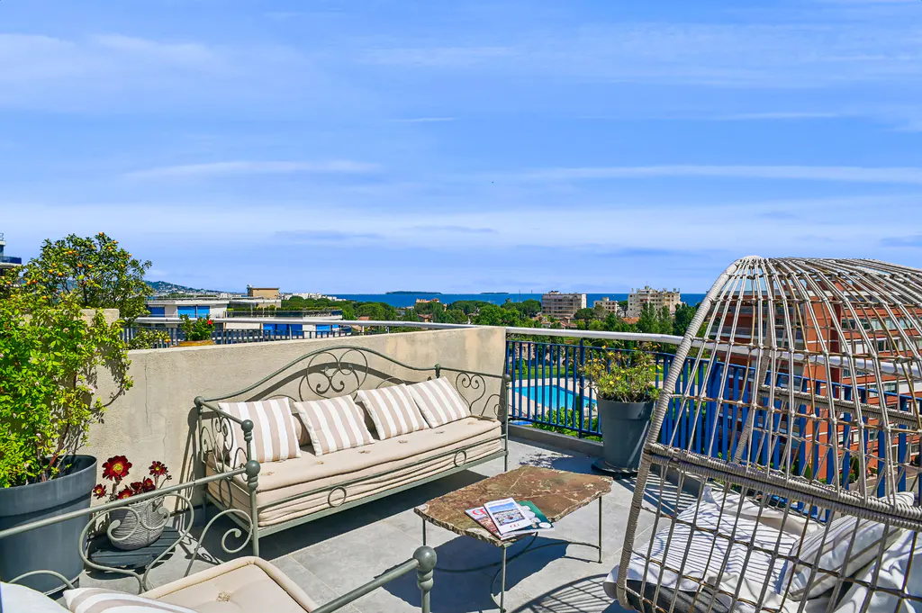 Rooftop patio with wrought iron furniture, including a sofa with striped pillows, a stone-topped table, and a hanging chair. City and ocean views.