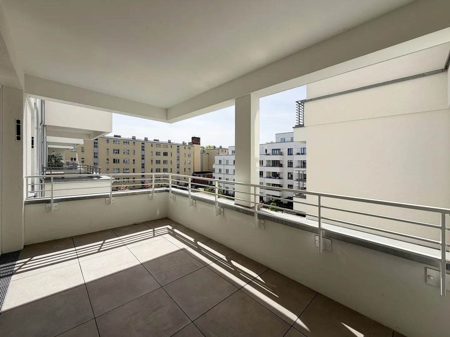 A bright, empty balcony with a tiled floor and white railings overlooks city buildings.