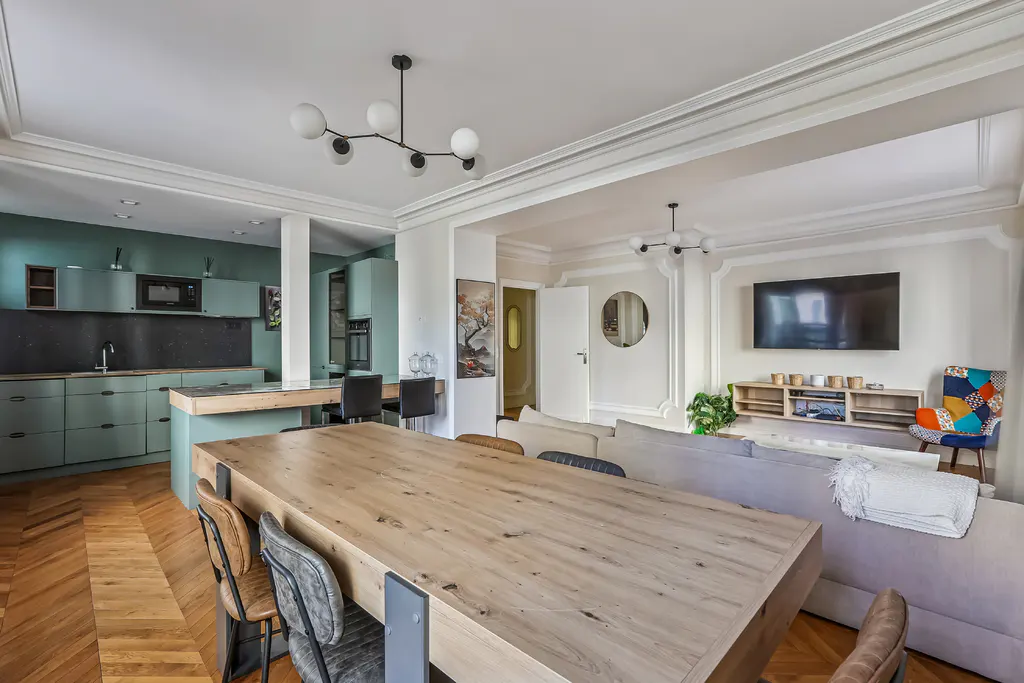 Open-concept living space with a large wooden table, herringbone floors, and a teal kitchen in the background. A gray sofa faces a wall-mounted TV.