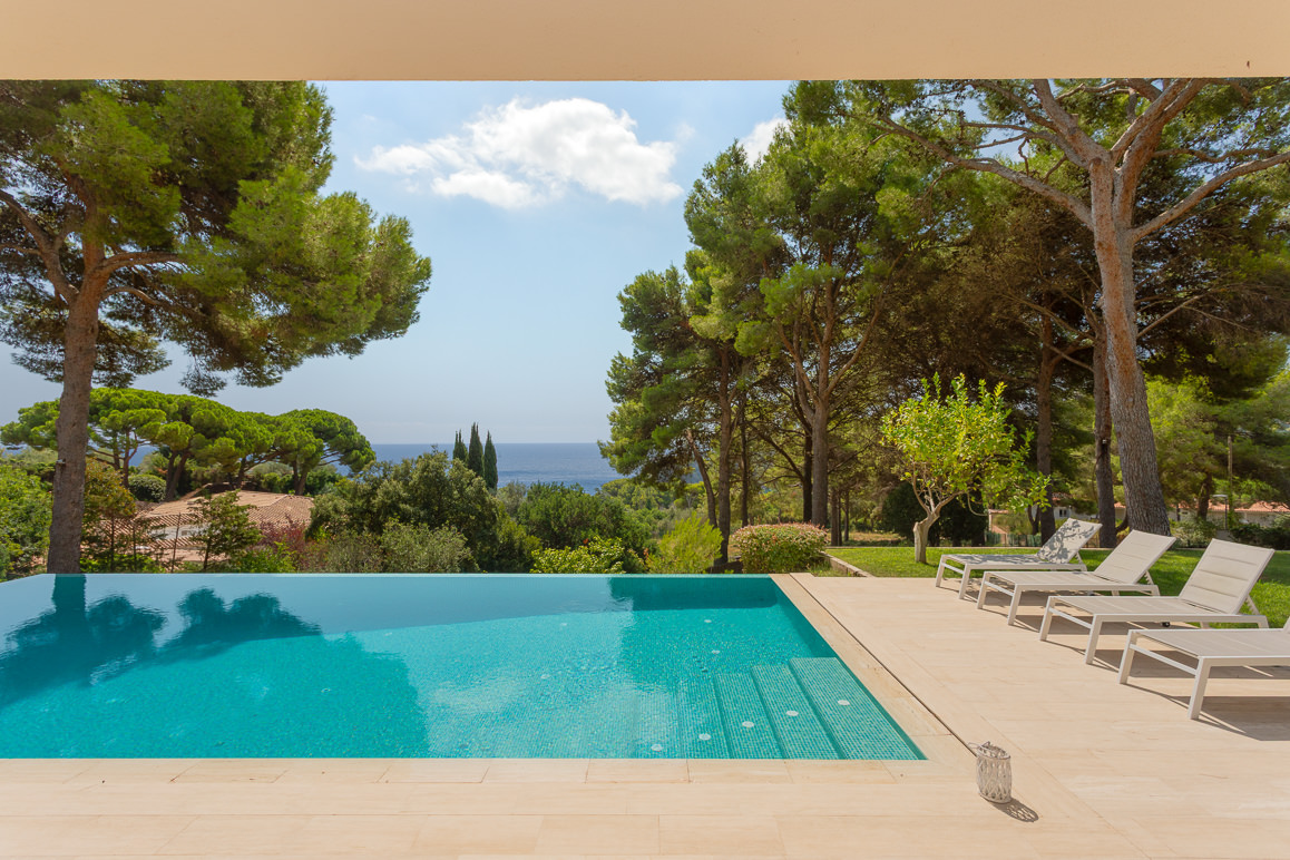 Infinity pool with turquoise water, stone deck, lounge chairs, and a scenic view of pine trees and the distant sea. [AI-generated text]