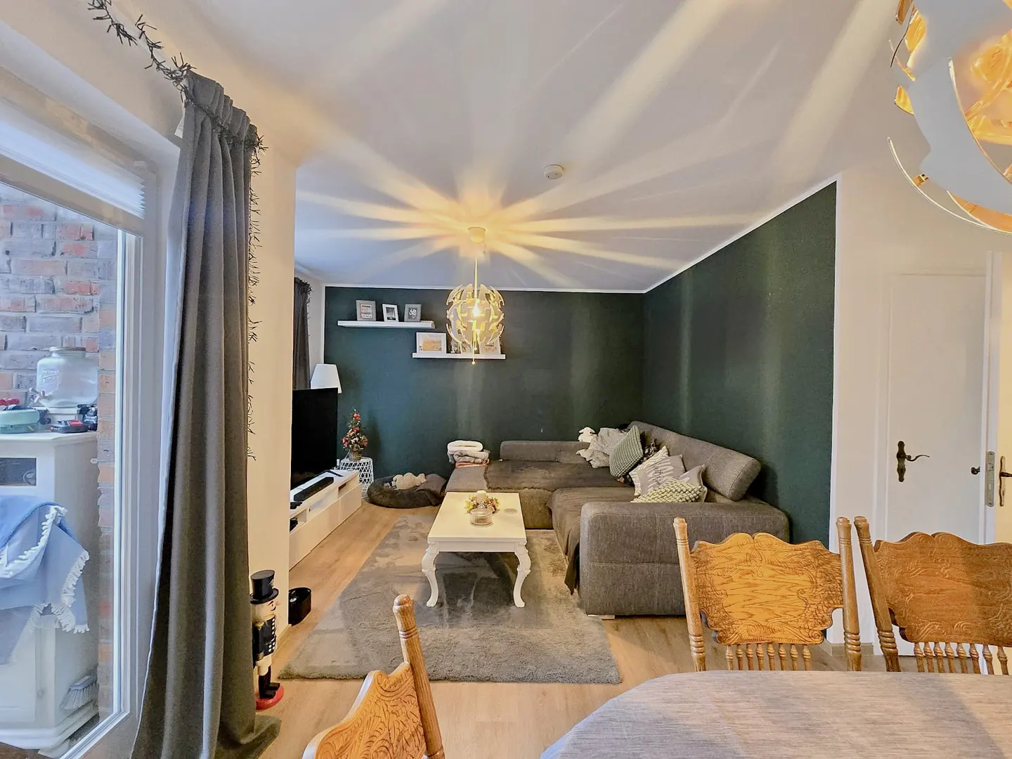 Living room with gray sectional sofa, white coffee table, and dark green accent wall. Wood chairs are in the foreground.