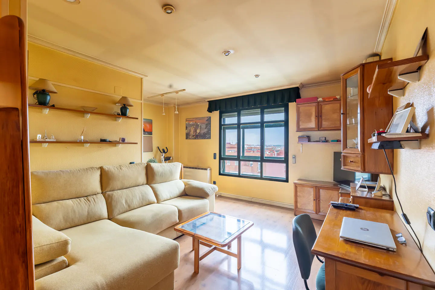 A living room with yellow walls, a beige sectional sofa, a wooden desk with a laptop, and a large window.