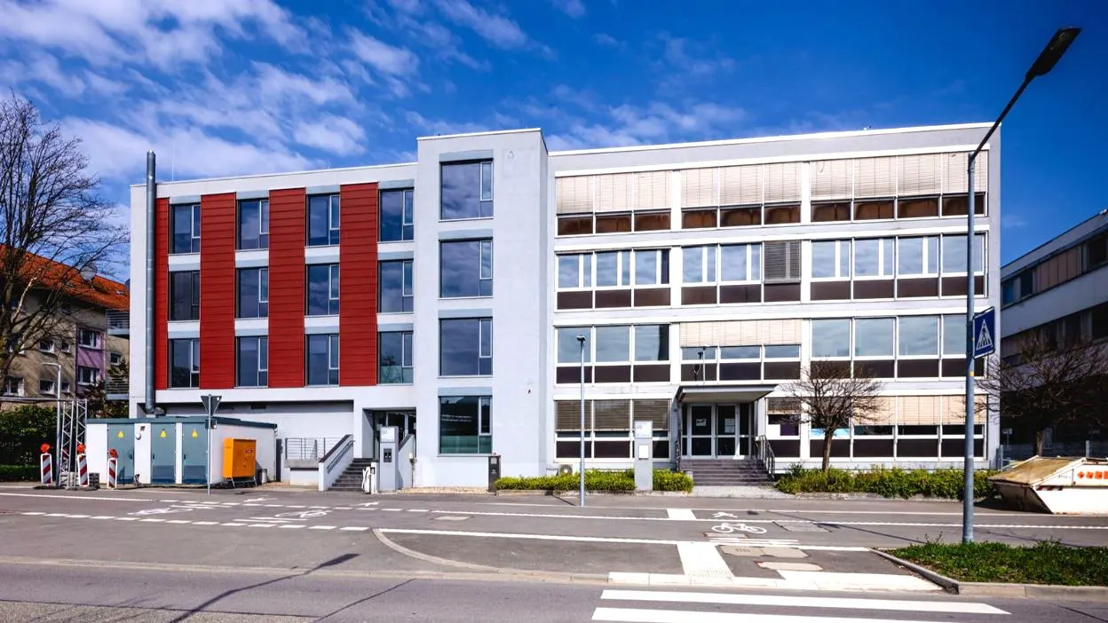 Modern, three-story building with red accents and large windows under a blue sky. A crosswalk is in the foreground.