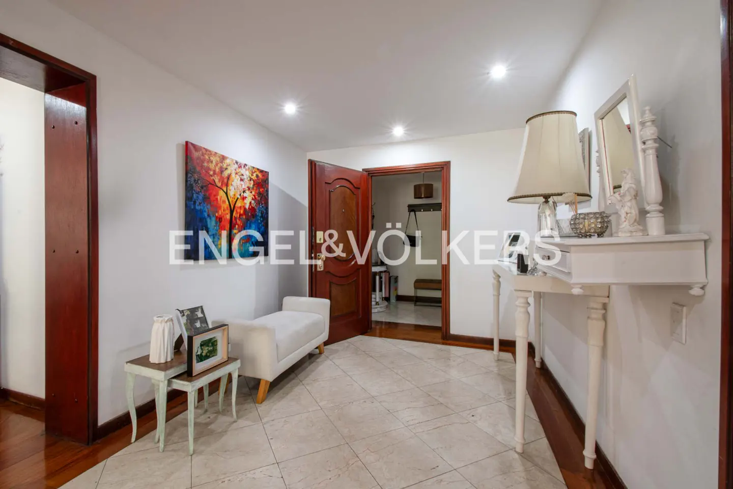 Bright foyer with marble floors, white walls, and recessed lighting. A white console table with a lamp and mirror stands near an open wood door. Artwork and a bench add color.
