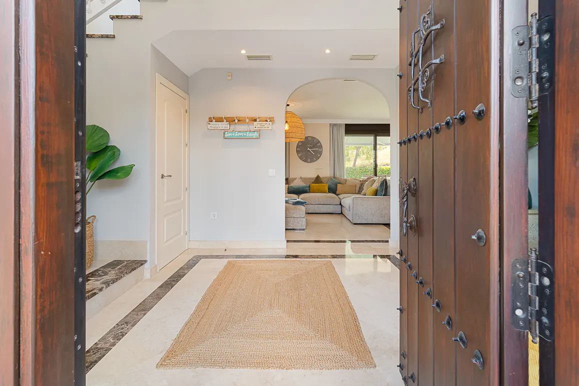 Open wooden door reveals a bright foyer with a jute rug, marble floors, and a glimpse of a living room with a large sofa.