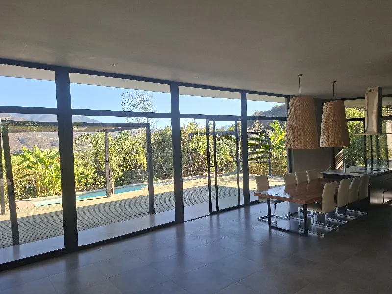 A modern dining room with a long wooden table and chairs, lit by two large woven pendant lights. Large windows offer a view of a pool and lush greenery.