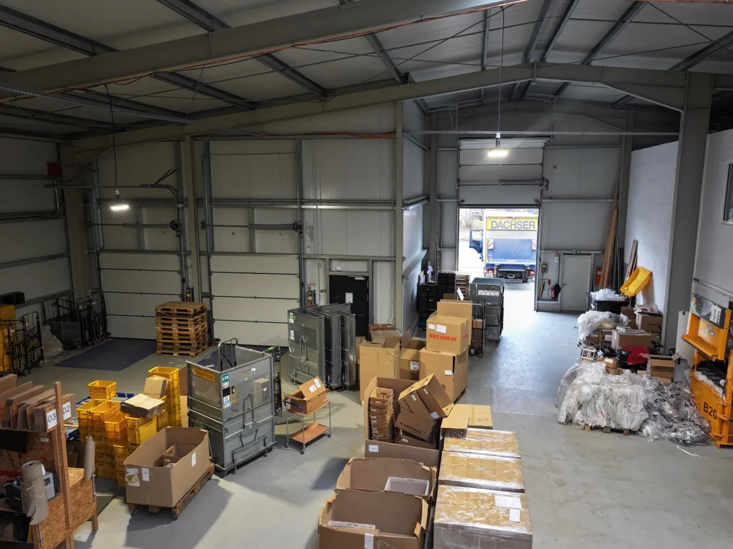 A high-angle view of a warehouse interior with boxes, pallets, and a truck visible through an open door.