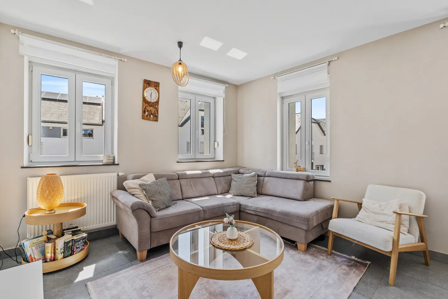 Bright living room with a gray sectional sofa, round glass coffee table, and white armchair on a light gray rug. Three windows with white frames.