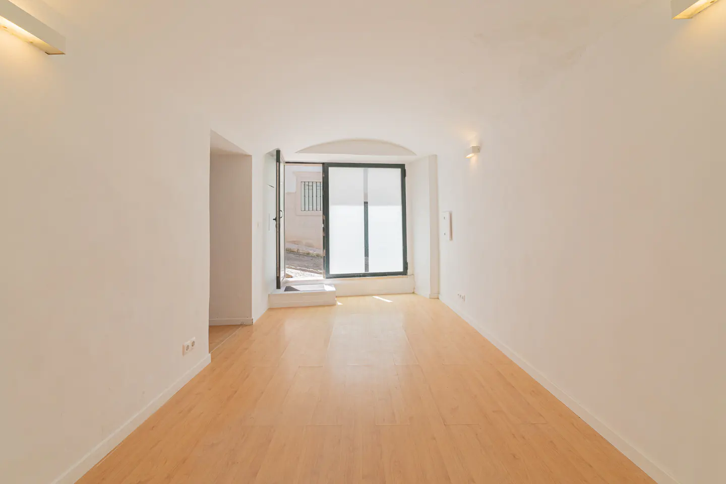 Bright, empty room with light wood floors and white walls. An open door leads to a street scene. Wall sconces provide soft lighting.