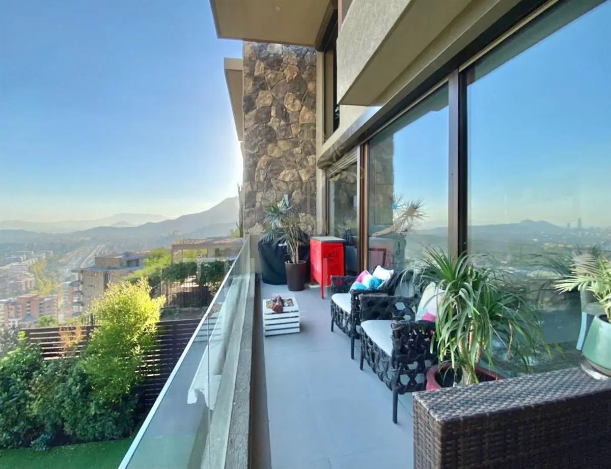 A modern balcony with wicker furniture, plants, and a stone wall overlooks a city and mountains on a sunny day.