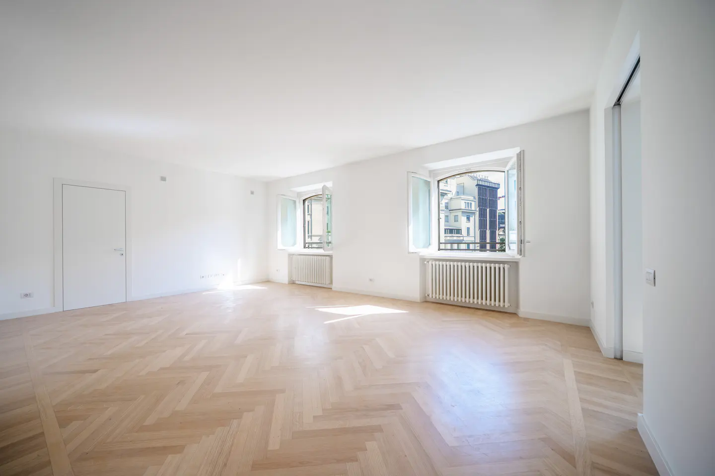 Bright, empty room with herringbone wood floors, white walls, two windows with radiators, and a closed white door.
