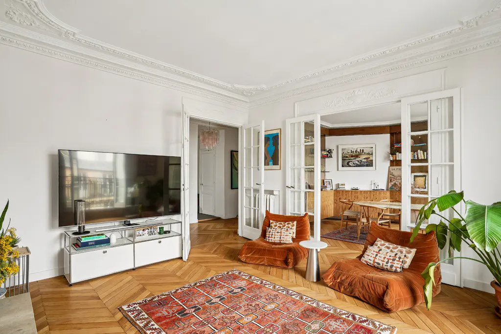 Bright living room with herringbone wood floors, white walls, and ornate ceiling trim. Two orange armchairs face a TV. A red rug sits in the center.