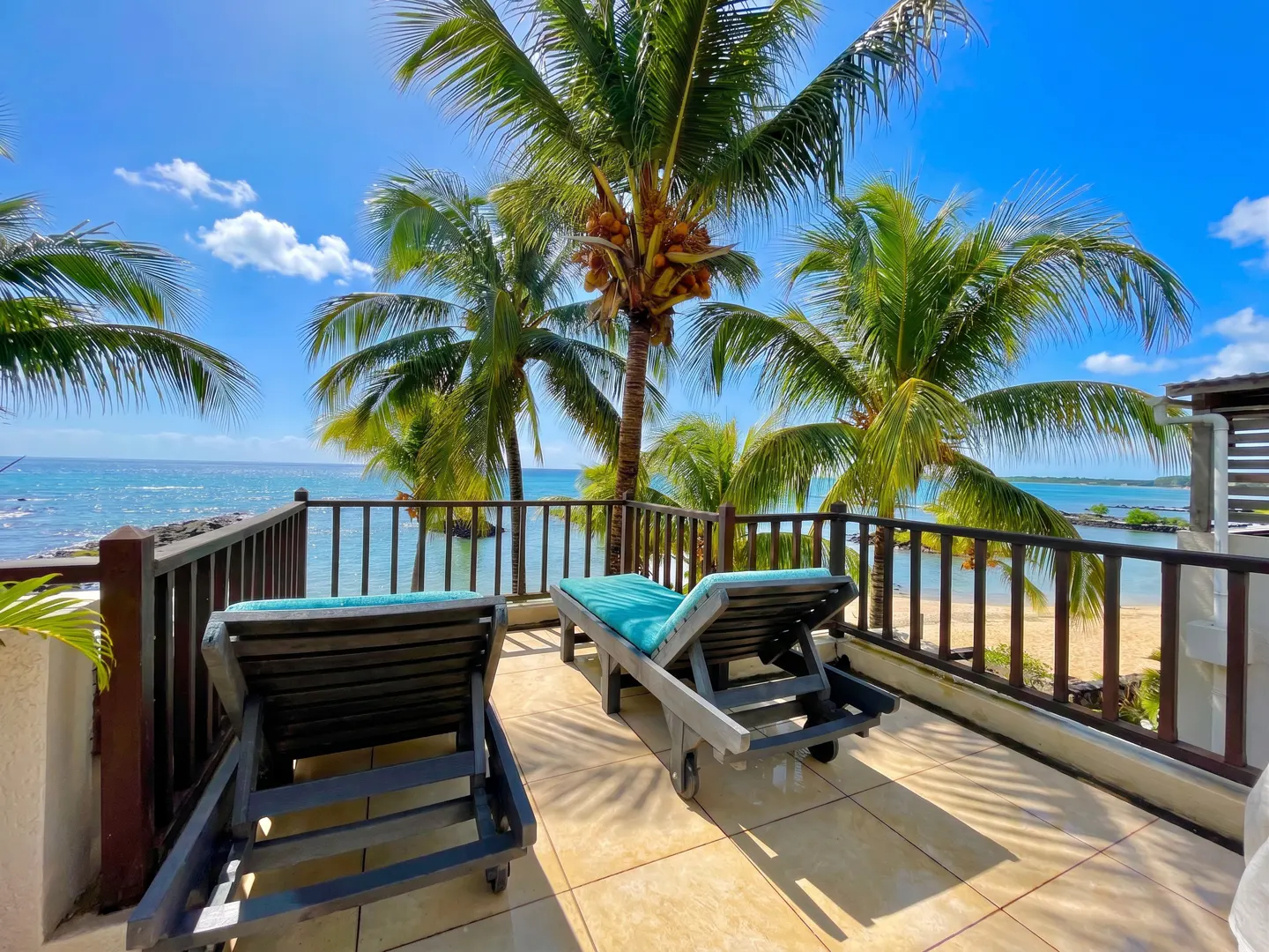 Balcony view with two lounge chairs, palm trees, and ocean. Blue cushions on gray chairs. Brown railing. Sunny day.